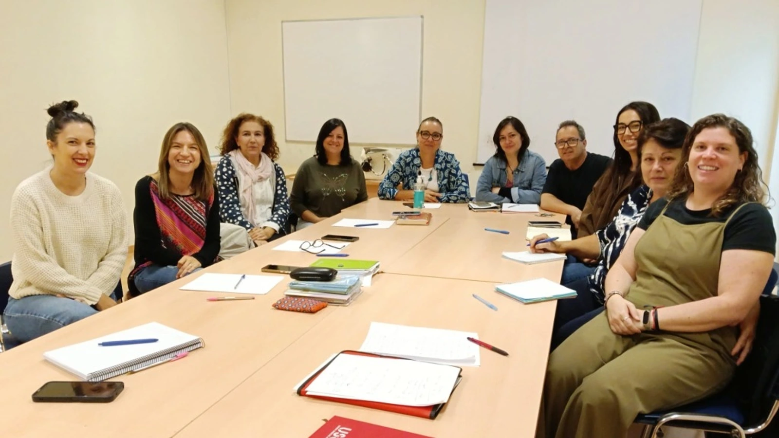 Varias personas sentadas alrededor de una mesa de reuniones con cuadernos y documentos en una sala con pizarra blanca