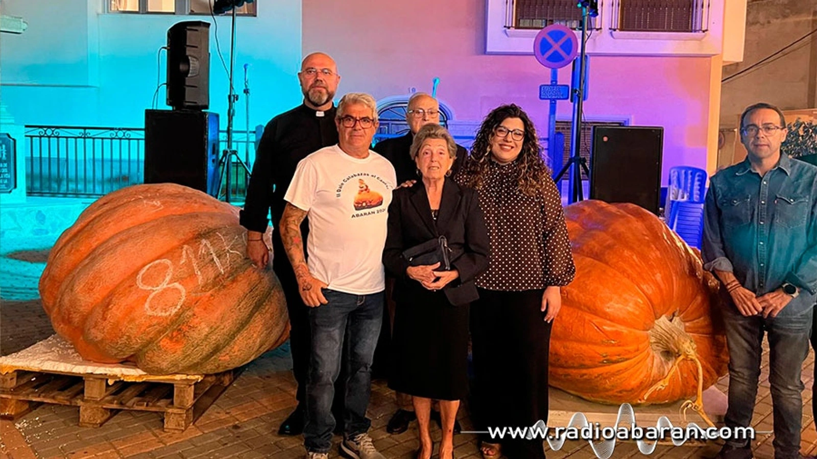 Dos calabazas gigantes junto a un grupo de personas, entre ellas un sacerdote, frente a un escenario iluminado en Abarán