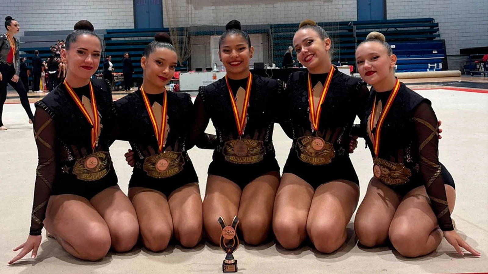 Cinco gimnastas con maillots negros del Club Cordillera posan arrodilladas sobre el tapiz con medallas de bronce y un trofeo de tercer puesto
