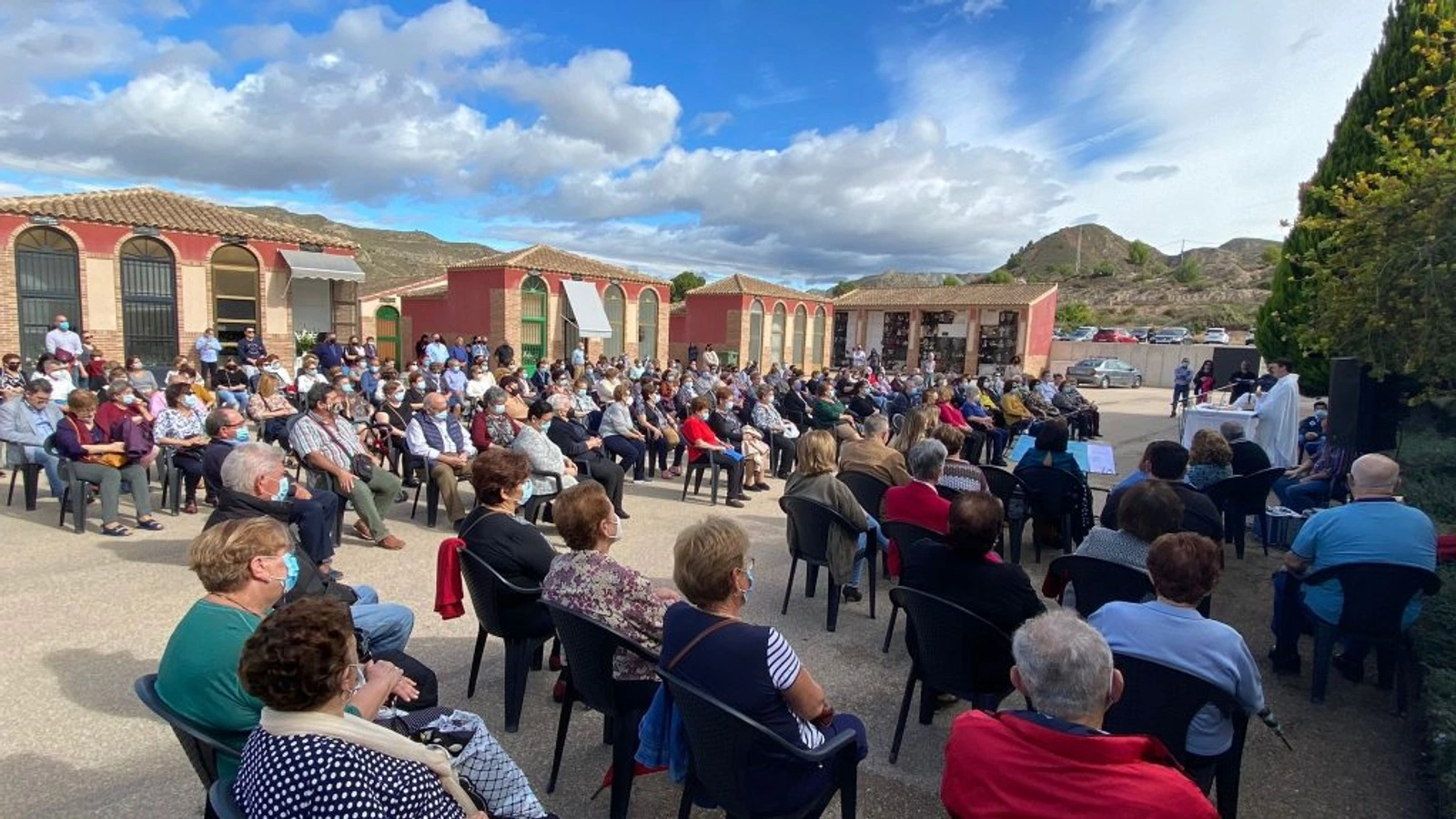 Decenas de vecinos sentados en sillas en la explanada del cementerio de Abarán durante una misa al aire libre, con bloques de nichos de ladrillo rojo al fondo y el sacerdote en el altar a la derecha.