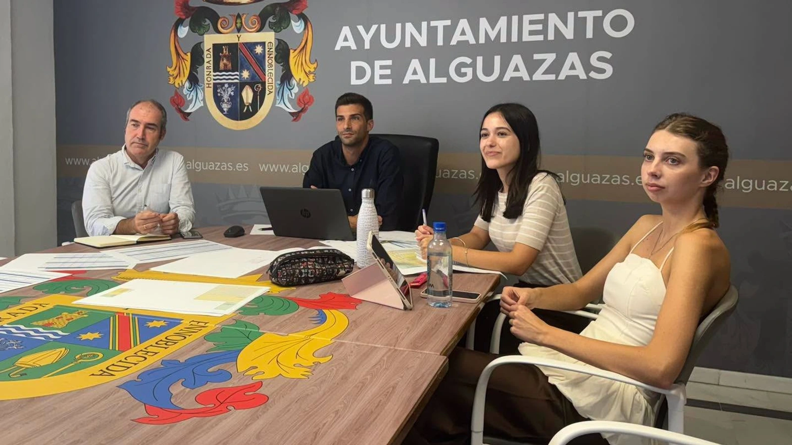 Cuatro personas en una mesa de reuniones con portátil y documentos ante el escudo del Ayuntamiento de Alguazas