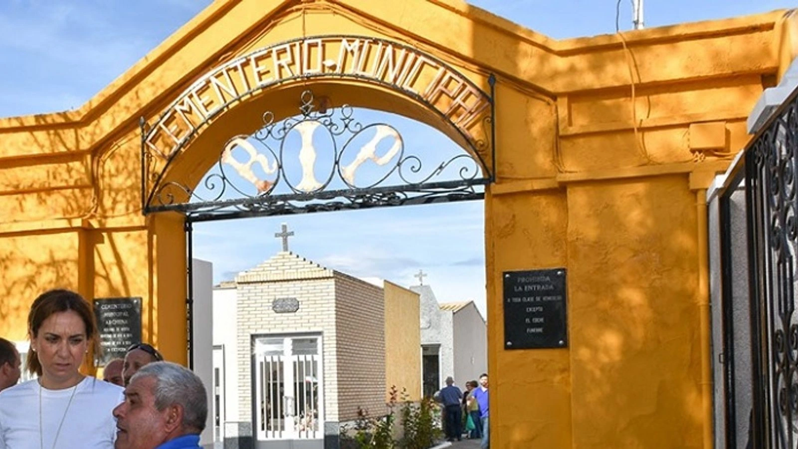 Portada amarilla de acceso al Cementerio Municipal de Archena, con arco de forja y rótulo, y varias personas en la entrada.