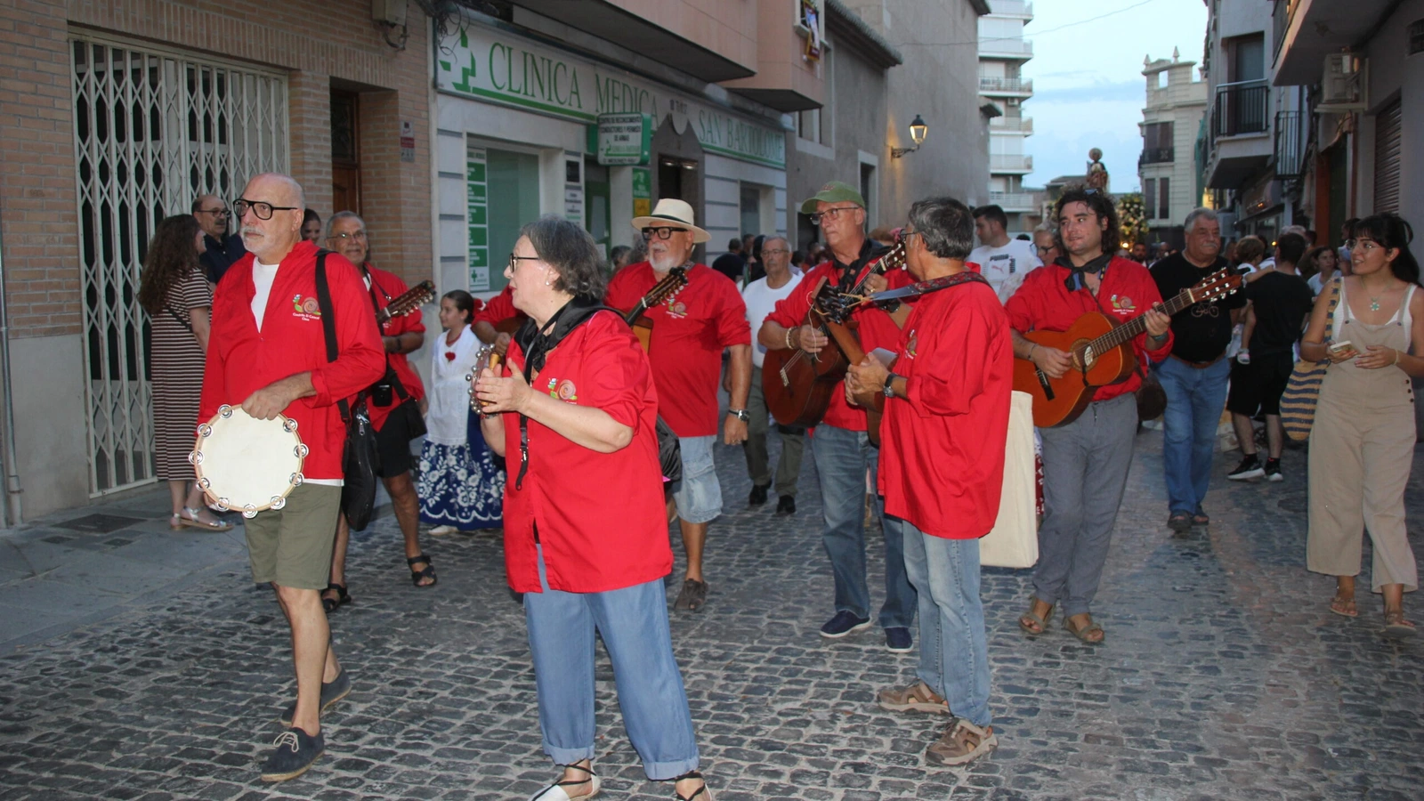 Músicos de la Cuadrilla El Caracol con camisas rojas tocan guitarras, laúdes y panderetas por una calle empedrada de Cieza