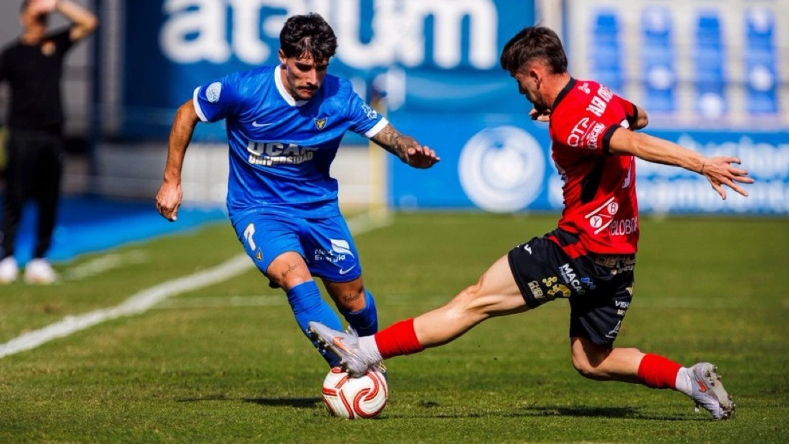 Un jugador del UCAM Murcia, de azul, conduce el balón ante la entrada de un futbolista del Cieza, de rojo y negro, en La Condomina