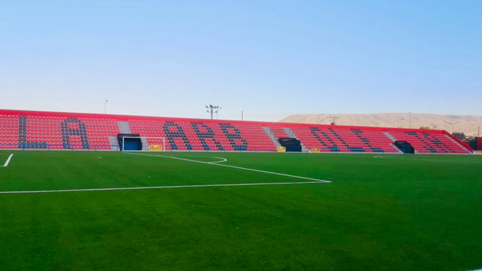 Graderío rojo con el nombre La Arboleja y el césped del estadio de Cieza, vacío