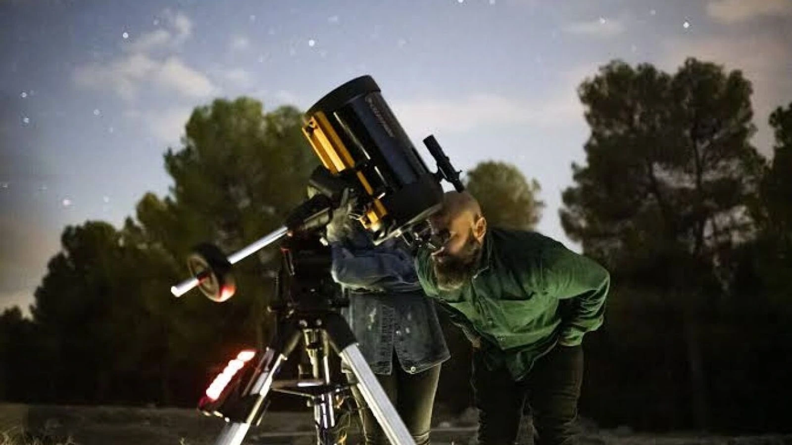 Dos personas observan el cielo por un telescopio de noche, junto a un trípode y entre pinos, bajo un cielo con estrellas