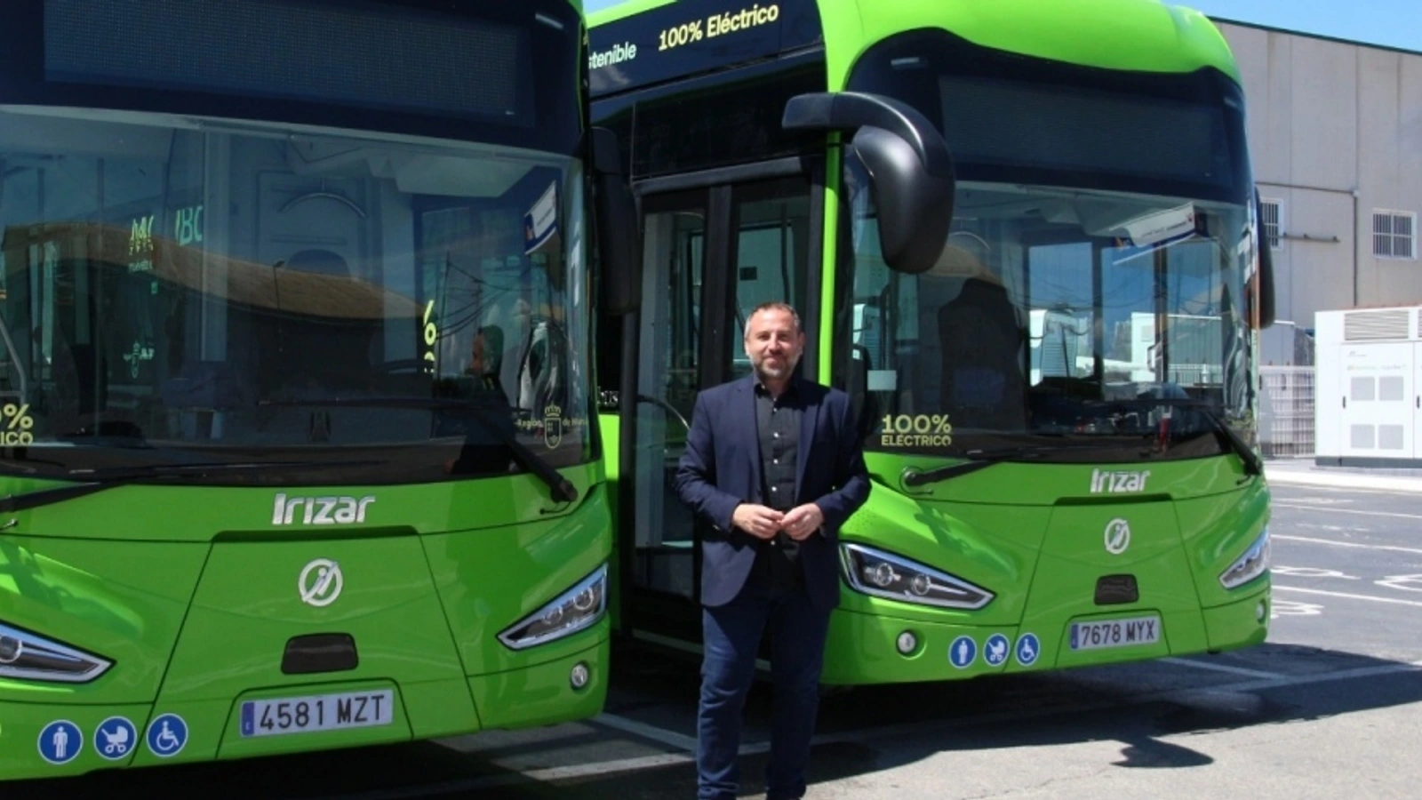 Un hombre con americana azul de pie entre dos autobuses verdes eléctricos aparcados
