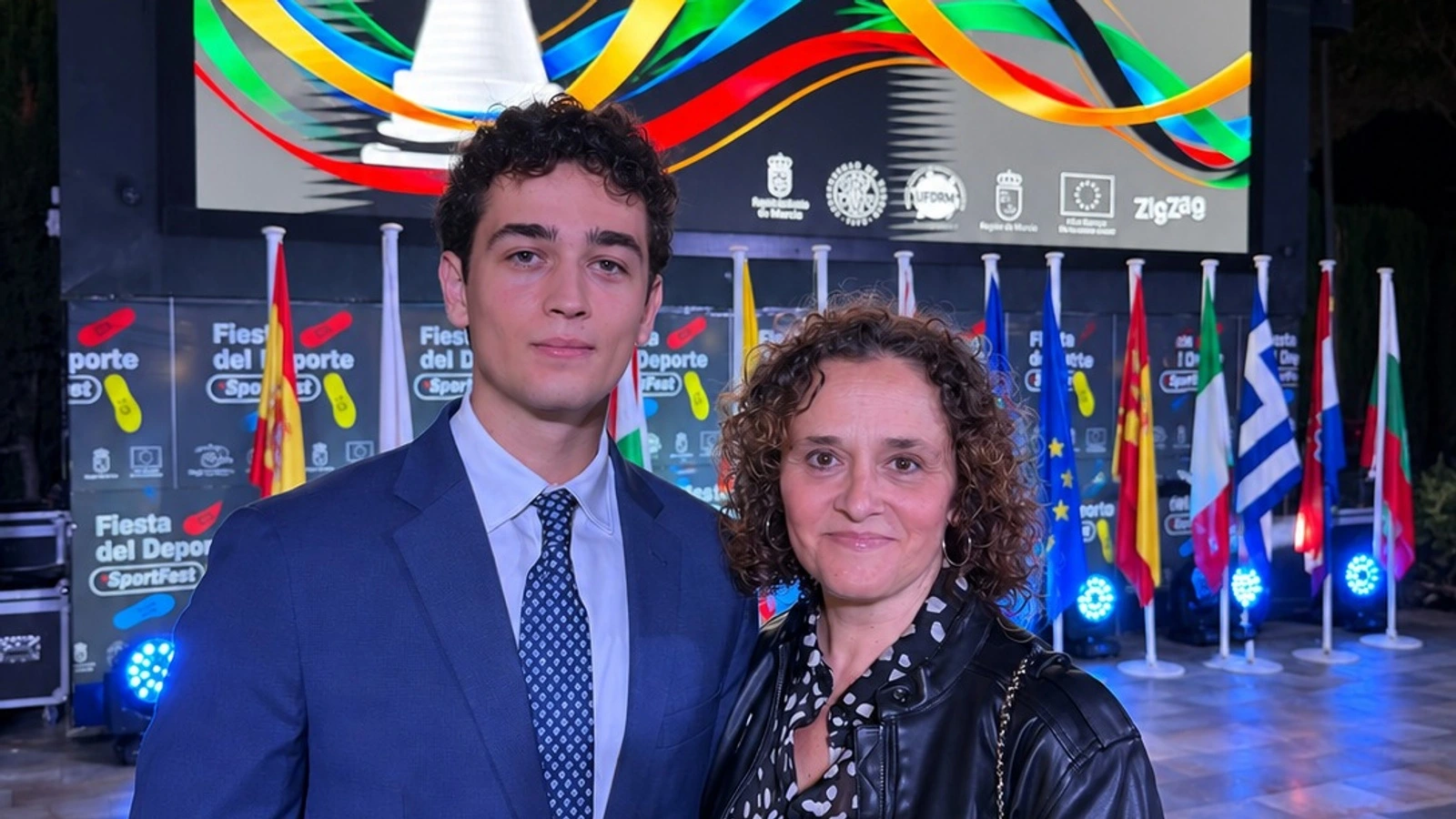 Ángel Turpín, con traje azul, posa junto a su madre ante la pantalla y las banderas de la V Gala del Deporte de la UFDRM en Murcia