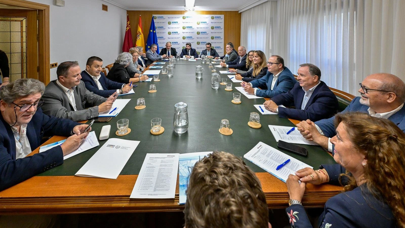 Una larga mesa de reuniones con una veintena de personas sentadas y un panel con el logotipo del Scrats al fondo