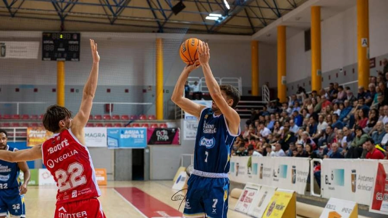 Un jugador de baloncesto con camiseta azul y el dorsal 7 lanza a canasta mientras un rival de rojo levanta el brazo