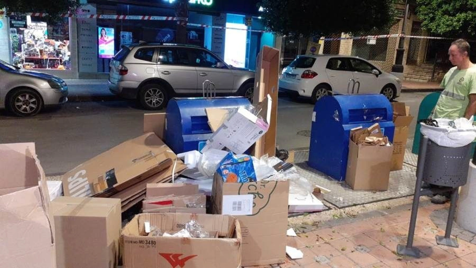 Cajas de cartón acumuladas junto a dos contenedores azules de papel en una calle de Molina de Segura, con un trabajador de Traperos al lado