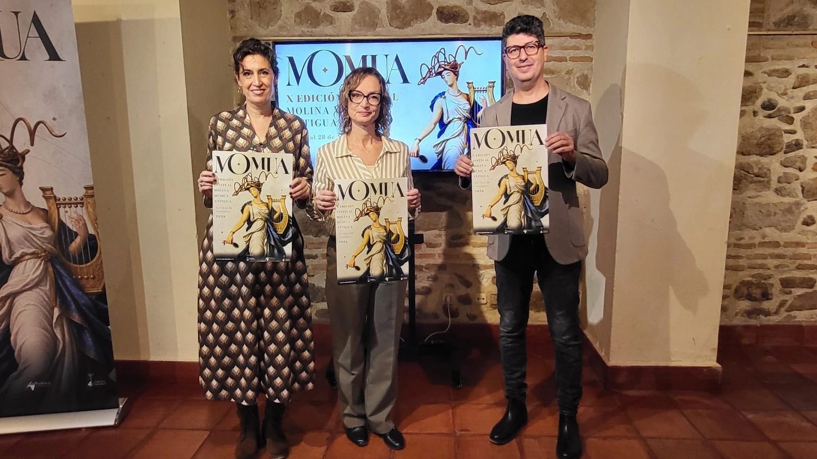 Dos mujeres y un hombre de pie sostienen carteles del festival MOMUA ante una pantalla y un muro de piedra