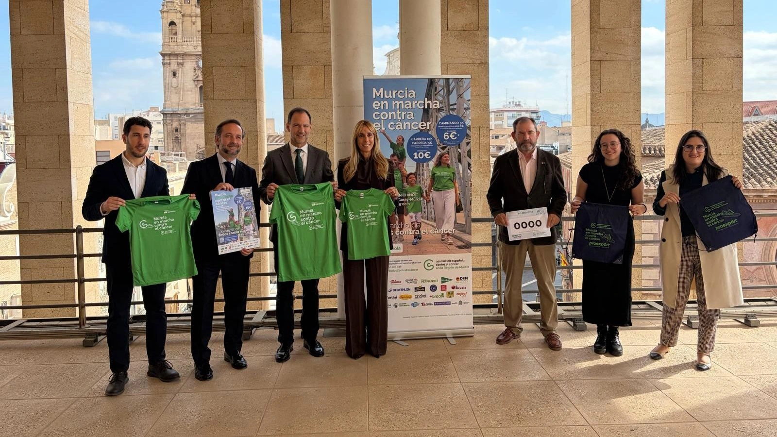 Siete personas sostienen camisetas verdes, mochilas y un dorsal de Murcia en Marcha contra el Cáncer junto al cartel de la prueba, con la torre de la Catedral de Murcia al fondo.