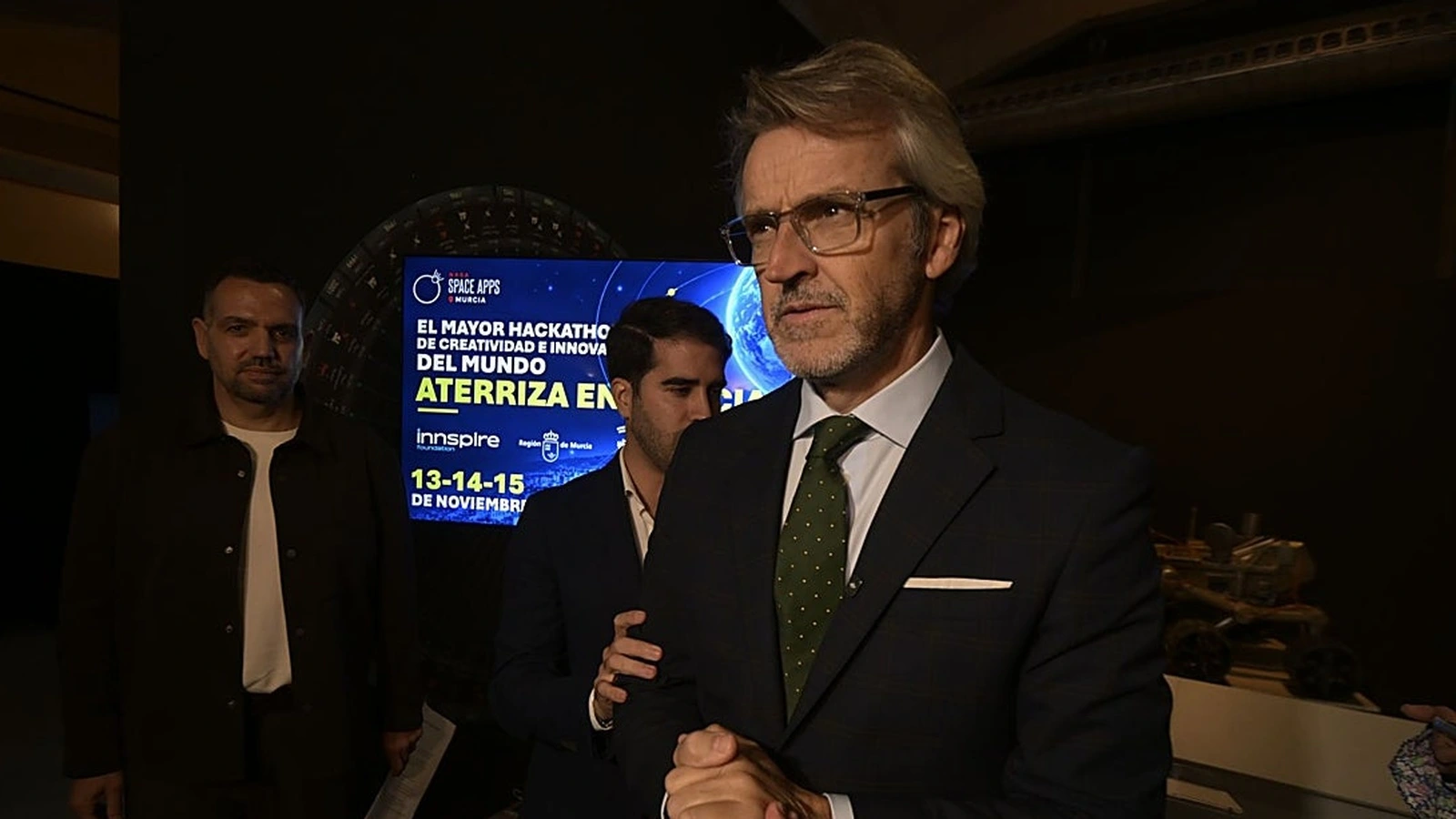 El consejero Luis Alberto Marín, con gafas y corbata verde, en primer plano ante una pantalla que anuncia el NASA Space Apps Murcia del 13 al 15 de noviembre