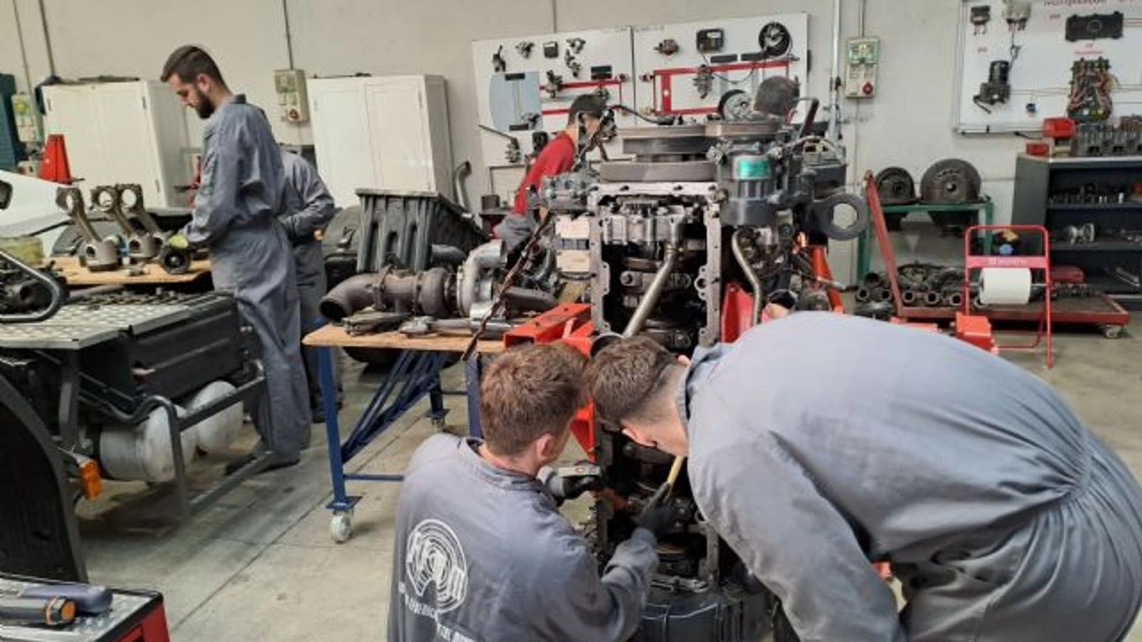 Alumnos con mono de trabajo reparan un motor en un taller durante un curso de formación profesional.