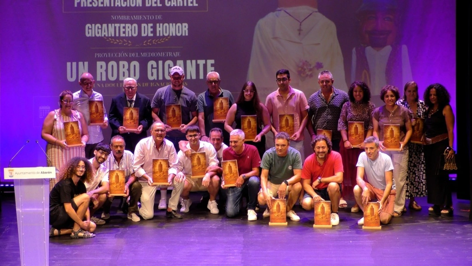 Grupo numeroso de personas posa en un escenario con placas conmemorativas ante una pantalla que anuncia Un robo gigante