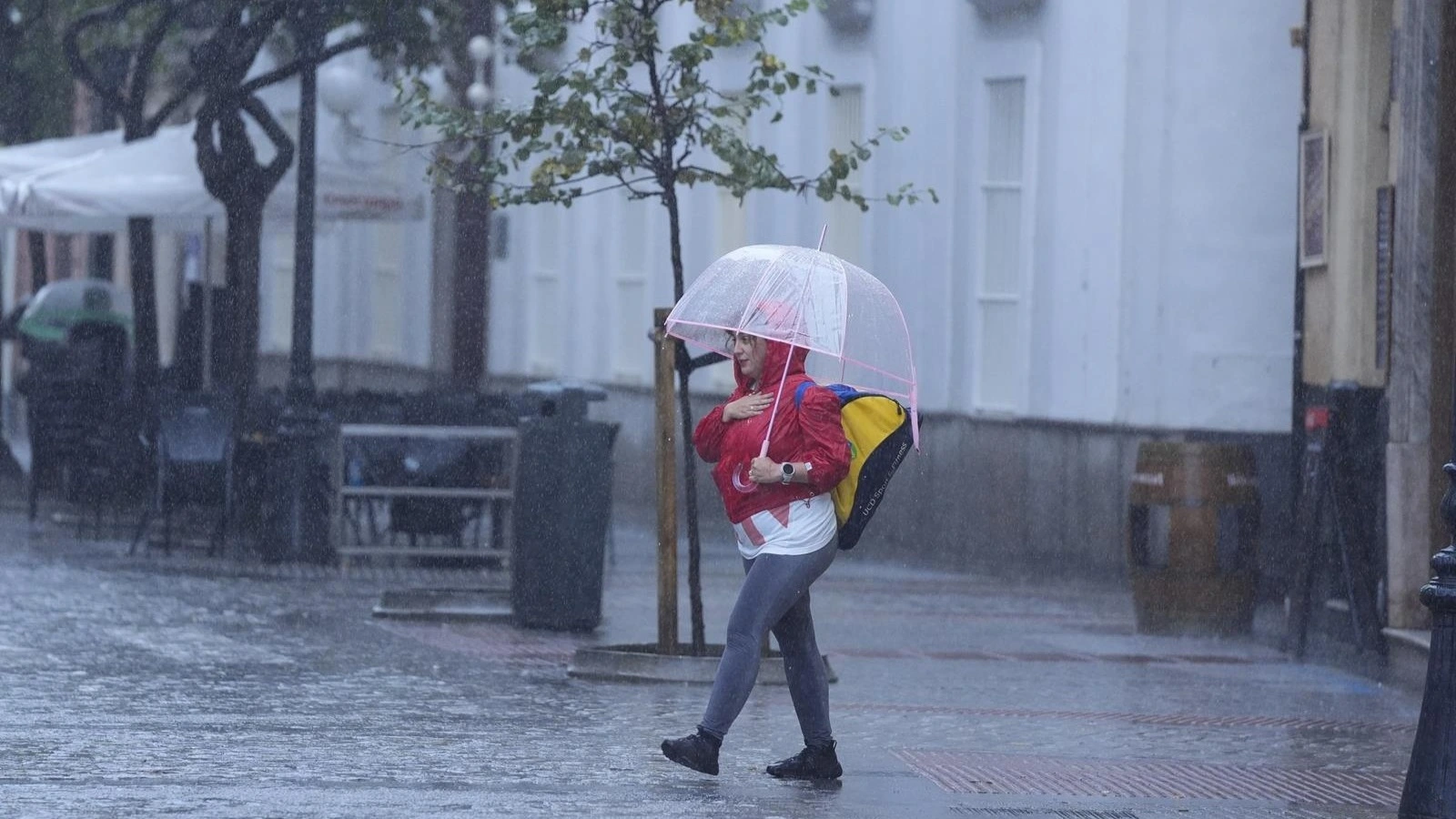 Una mujer con chubasquero rojo y paraguas transparente cruza una calle mojada bajo la lluvia